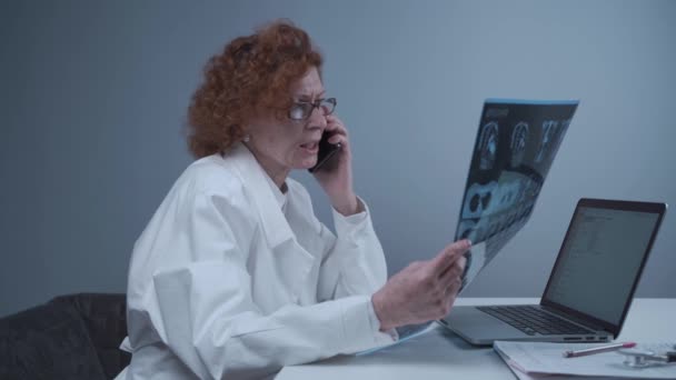 Female doctor looking at CT x-ray talking to colleague on smart phone in hospital office. Doctor on the phone in clinic, examining CT scans and discussing his findings. Radiologist lung x-ray exam — Vídeo de Stock