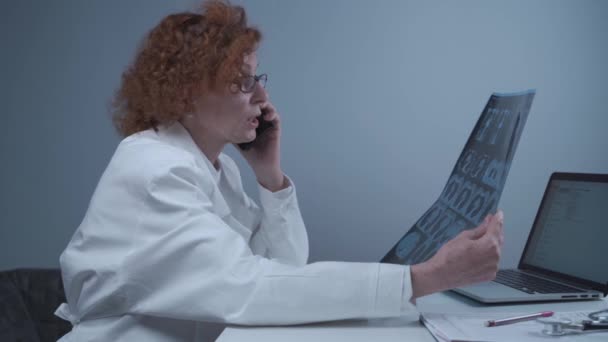 Счастливая взрослая женщина врач-рентгенолог обсуждает компьютерную томографию легких с коллегами по телефону в больничном кабинете. Пулмонолог изучает МРТ или пульмонографию и использует сотовый телефон. Рентген грудной клетки — стоковое видео