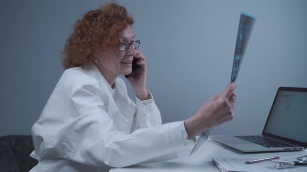 Anciana caucásica mujer médico analizar la imagen de rayos X y hablar con el teléfono móvil acerca de los diagnósticos en el consultorio médico. Médico especialista que trabaja con la tomografía computarizada, hable con el teléfono celular sobre los resultados de la radiografía — Vídeo de stock