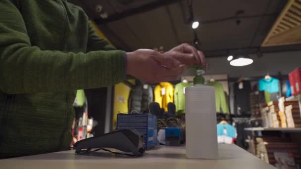 Salesperson at sports store uses hand sanitizer while serving customer. Seller at travel store uses an alcohol gel behind counter. Man washing hands with antiseptic at shop counter. New norm covid 19 — Vídeo de Stock