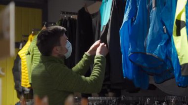 Masked man in sports and outdoor equipment store selects jacket during covid 19 quarantine and keeps social distance. A male buyer in a mask buys sportswear for trekking and hiking outdoors