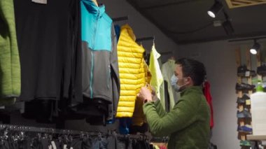 Man in protective mask shopping outdoor equipment in sports store during quarantine covid 19. Masked male looking to buy jacket in tourist shop. Shopper choosing sportswear in sporting goods store