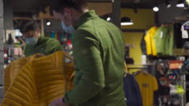 Topic of buying sportswear in offline sports store during new norms pandemic for covid 19. Man wearing mask chooses jacket in an outdoor store. Masked male looking to buy jacket during shopping
