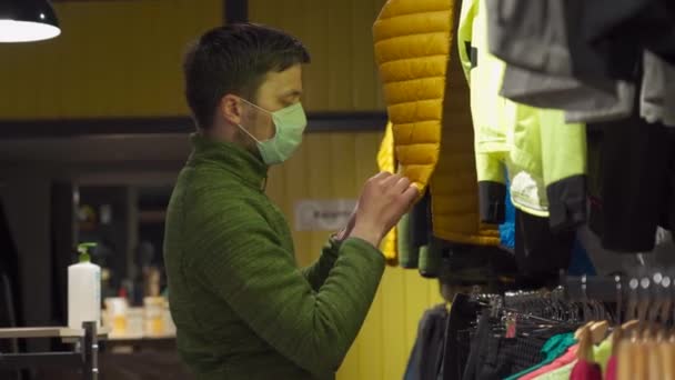 El hombre enmascarado en la tienda de deportes y equipos al aire libre selecciona la chaqueta durante la cuarentena covid 19 y mantiene la distancia social. Un comprador masculino en una máscara compra ropa deportiva para trekking y senderismo al aire libre — Vídeos de Stock