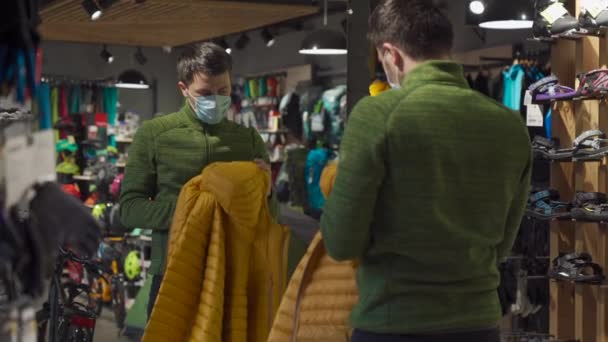 Man in protective mask shopping outdoor equipment in sports store during quarantine covid 19. Masked male looking to buy jacket in tourist shop. Shopper choosing sportswear in sporting goods store — Wideo stockowe