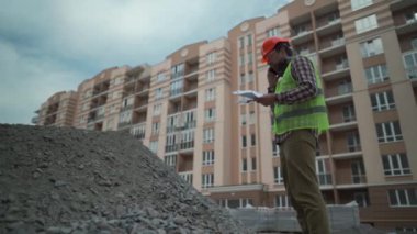 Satisfied foreman in helmet and vest at construction site talking on phone with rubble contractor and looks at waybills. Builder will check arrival building material, check shortage and call by phone