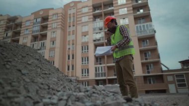 Construction worker angry with cell phone and checklist control imported building material by contractors. Foreman in protective overalls at site with documents and phone stands near heap of rubble