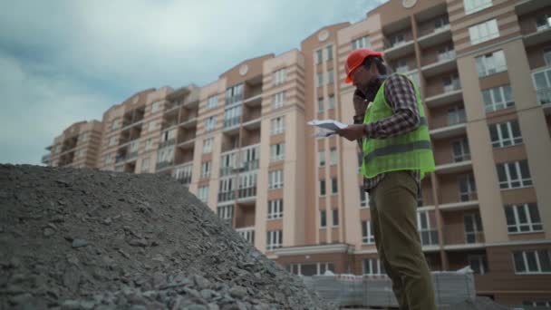 Satisfied foreman in helmet and vest at construction site talking on phone with rubble contractor and looks at waybills. Builder will check arrival building material, check shortage and call by phone — Wideo stockowe