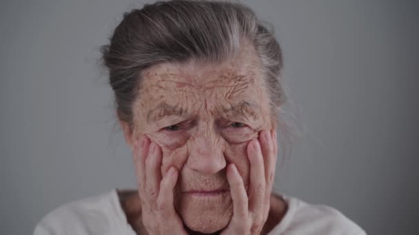 Старша жінка 90 років з глибокими зморшками і сірим волоссям має психологічний стрес, плаче і дивиться на камеру, що тримає її обличчя руками на сірому фоні. Депресія і самотність у літніх людей — стокове відео