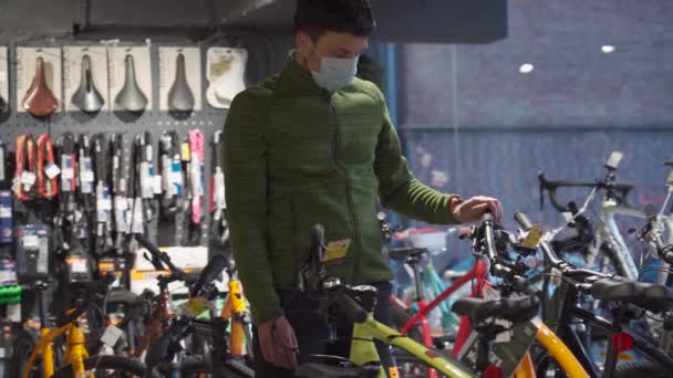 Shopper i steril mask välja mountainbikes i sportaffär. Mannen som bär skyddsmask väljer cykel i cykelaffär med socialt avstånd under coronavirus karantän. Ny norm — Stockvideo