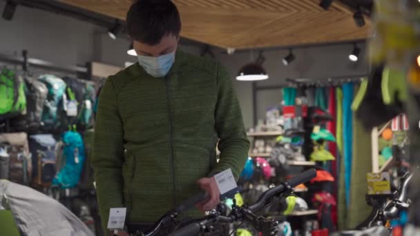 Indecisive masked man at bicycle store chooses sports bike from an assortment. Customer in bicycle shop, looking at price tag during difficult choice from large assortment of bicycles in sports shop — Stock Video