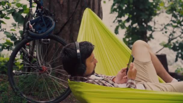 Ung kaukasisk man vilar i hängmatta, lyssnar på musik på hörlurar och använder smartphone efteråt på cykel i skogen nära sjön. Ljud läkning. Idylliskt ställe. Resor, camping i naturen — Stockvideo