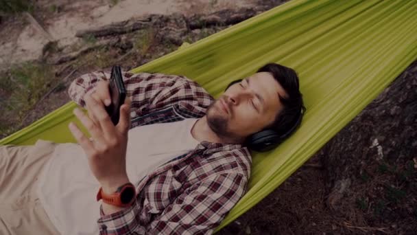 O homem viaja de bicicleta, relaxando em rede verde, navegando na Internet no smartphone, ouvindo música em fones de ouvido na floresta perto do lago. Ciclista em rede no acampamento por rio. Masculino de bicicleta na rede — Vídeo de Stock