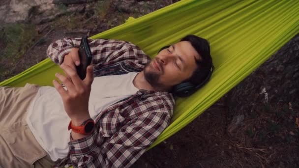 Homem em viagem de bicicleta no acampamento junto ao lago está relaxando em rede verde enquanto ouve música. Tema de recreação ativa na natureza. Hipster ciclista com fones de ouvido se divertindo em rede por rio — Vídeo de Stock