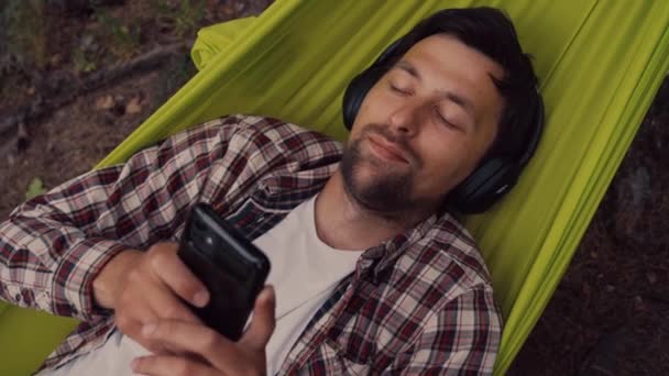 Jovem caucasiano descansando na rede, ouvindo música em fones de ouvido e usando smartphone depois em bicicleta na floresta perto do lago. Recuperação de áudio. Lugar idílico. Viajar, acampar na natureza — Vídeo de Stock