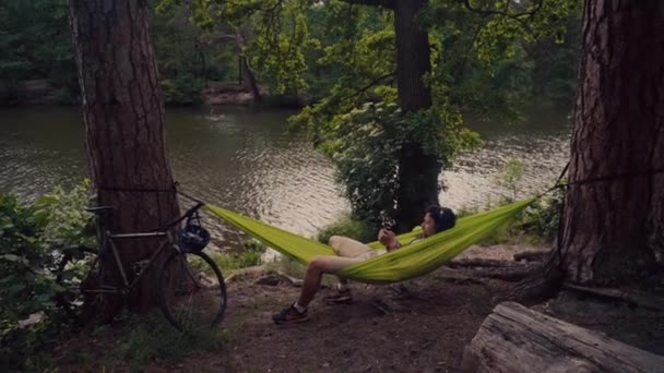 Människan färdas på cykel, koppla av i grön hängmatta, surfa på Internet på smartphone, lyssna musik på hörlurar i skogen nära sjön. Cyklist i hängmatta vid campingen vid ån. Man på cykel i hängmatta — Stockvideo