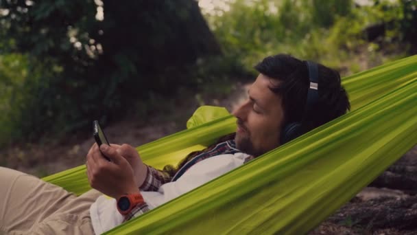 El hombre en viaje de la bici en acampar por el lago está relajando en hamaca verde mientras que escucha música. Tema de recreación activa en la naturaleza. Hipster ciclista con auriculares divertirse en hamaca por río — Vídeo de stock