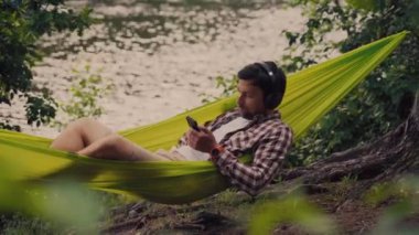 Genç beyaz adam hamakta dinleniyor, kulaklıkla müzik dinliyor ve sonrasında gölün yanındaki ormanda bisiklette akıllı telefon kullanıyor. Ses iyileştirme. Idyllic bir yer. Seyahat, doğada kamp.