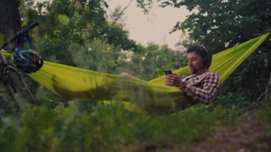 Göl kenarındaki kampta bisiklet süren bir adam müzik dinlerken yeşil bir hamakta dinleniyor. Doğada aktif eğlence teması. Kulaklıklı hippi bisikletçi nehir kenarındaki hamakta eğleniyor.