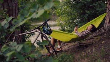 Açık hava ve macera konsepti. Ormana bisiklet gezisi. Bisikletçi, kulaklıkla müzik dinlerken ve akıllı telefonda internette sörf yaparken doğadaki ağaçlar arasındaki yeşil hamakta dinleniyor.