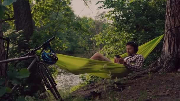 Giovane uomo caucasico che riposa in amaca, ascoltando musica sulle cuffie e utilizzando lo smartphone in seguito in bicicletta nella foresta vicino al lago. Guarigione audio. Un posto idilliaco. Viaggi, campeggio nella natura — Video Stock