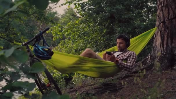 Man travels on bicycle, relaxing in green hammock, surfing Internet on smartphone, listening music on headphones in forest near lake. Cyclist in hammock at campsite by river. Male on bike in hammock — Stock Video