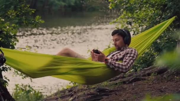 Al aire libre y concepto de aventura. Viaje en bicicleta al bosque. ciclista está descansando en hamaca verde entre los árboles en la naturaleza junto al lago mientras escucha música en los auriculares y navegar por Internet en el teléfono inteligente — Vídeo de stock