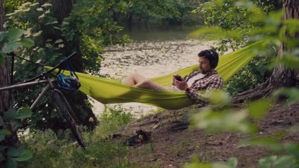 Man travels on bicycle, relaxing in green hammock, surfing Internet on smartphone, listening music on headphones in forest near lake. Cyclist in hammock at campsite by river. Male on bike in hammock — Stock Video