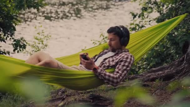 Homem em viagem de bicicleta no acampamento junto ao lago está relaxando em rede verde enquanto ouve música. Tema de recreação ativa na natureza. Hipster ciclista com fones de ouvido se divertindo em rede por rio — Vídeo de Stock