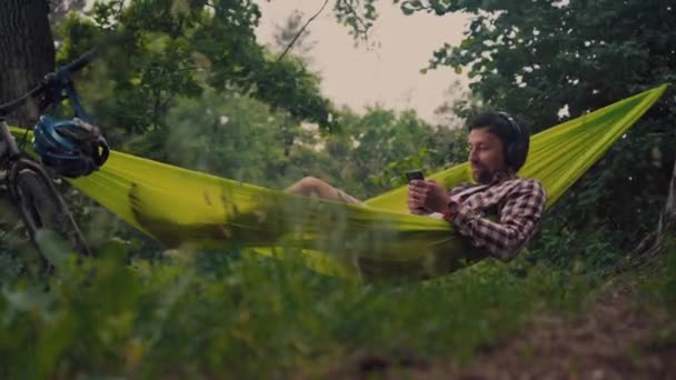 Man travels on bicycle, relaxing in green hammock, surfing Internet on smartphone, listening music on headphones in forest near lake. Cyclist in hammock at campsite by river. Male on bike in hammock — Stock Video