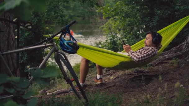 Man på cykeltur på camping vid sjön är avkopplande i grön hängmatta medan du lyssnar på musik. Aktivt fritidstema i naturen. Hipster cyklist med hörlurar som har kul i hängmatta vid flod — Stockvideo
