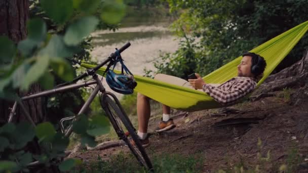 Ao ar livre e conceito de aventura. Viagem de bicicleta para a floresta. Ciclista está descansando em rede verde entre árvores na natureza por lago enquanto ouve música em fones de ouvido e navegar na Internet no smartphone — Vídeo de Stock