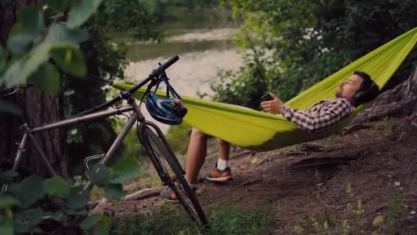 O homem viaja de bicicleta, relaxando em rede verde, navegando na Internet no smartphone, ouvindo música em fones de ouvido na floresta perto do lago. Ciclista em rede no acampamento por rio. Masculino de bicicleta na rede — Vídeo de Stock