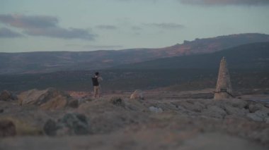 Sırt çantalı genç adam gün batımında Akdeniz adasında Akdeniz kıyısında kayalık sahil boyunca yürürken akıllı telefondan fotoğraf çekiyor. Sırt çantalı gezgin güzel deniz manzarasının fotoğraflarını çeker.