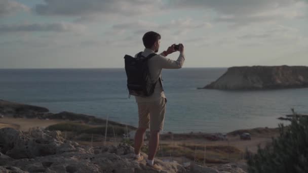 Sırt çantalı genç adam gün batımında Akdeniz adasında Akdeniz kıyısında kayalık sahil boyunca yürürken akıllı telefondan fotoğraf çekiyor. Sırt çantalı gezgin güzel deniz manzarasının fotoğraflarını çeker. — Stok video