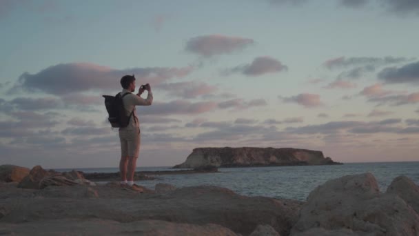 Toerist maakt foto 's van een prachtig uitzicht op de natuur op het eiland Cyprus op mobiele telefoon tijdens het wandelen langs rotsachtige kust Middellandse Zee bij zonsondergang. Man op rotsachtige kustlijn neemt foto 's met de smartphone — Stockvideo