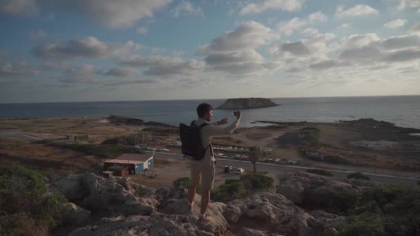 Resenären upprätthåller resor blogg ta bilder och videor med smartphone medan promenader längs steniga kusten havet på ön cyprus vid solnedgången. Backpacker gör foto havsutsikt på stenig strand på telefon — Stockvideo