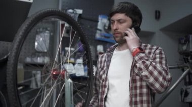 Store employee takes order by mobile phone standing in bicycle workshop near cycle wheel. Bike shop owner at work. Mechanic with clipboard talking on mobile phone. Guy calls after fixing bike