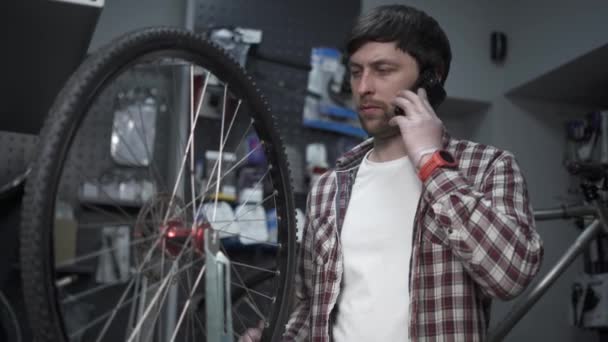 Store employee takes order by mobile phone standing in bicycle workshop near cycle wheel. Bike shop owner at work. Mechanic with clipboard talking on mobile phone. Guy calls after fixing bike — ストック動画