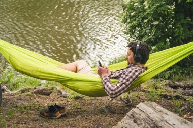 Genç beyaz adam hamakta dinleniyor, kulaklıkla müzik dinliyor ve sonrasında gölün yanındaki ormanda bisiklette akıllı telefon kullanıyor. Ses iyileştirme. Idyllic bir yer. Seyahat, doğada kamp..