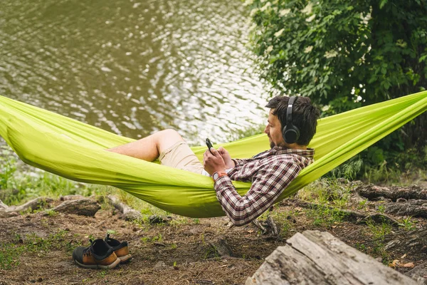 屋外と冒険の概念 森への自転車旅行 サイクリストは ヘッドフォンで音楽を聴いたり スマートフォンでインターネットサーフィンをしたりしながら 湖で自然の木々の間に緑のハンモックで休んでいます — ストック写真