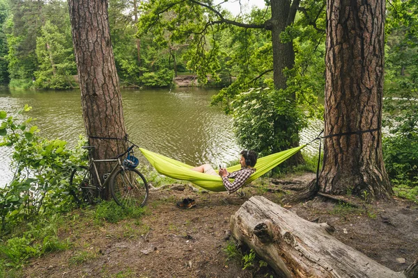 湖でのキャンプでの自転車旅行の男は音楽を聴きながら緑のハンモックでリラックスしています 自然の中でアクティブなレクリエーションのテーマ ヒップスターサイクリストとともにヘッドフォン楽しみを持っていますハンモックで川 — ストック写真