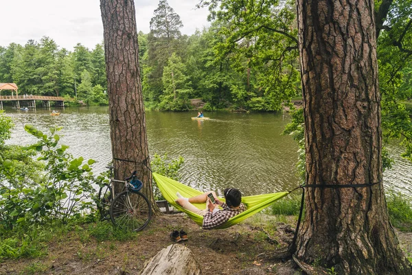 ハンモックに寝そべってヘッドフォンで音楽を聴いたり 湖の近くの森の中の自転車でスマートフォンを使用している若い白人男性 オーディオヒーリング 牧歌的な場所だ 自然の中でキャンプ — ストック写真