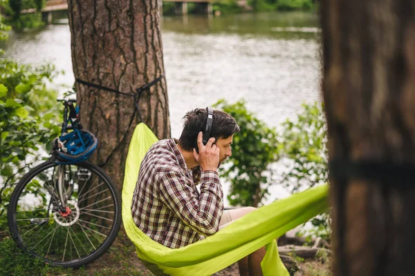 屋外と冒険の概念 森への自転車旅行 サイクリストは ヘッドフォンで音楽を聴いたり スマートフォンでインターネットサーフィンをしたりしながら 湖で自然の木々の間に緑のハンモックで休んでいます — ストック写真