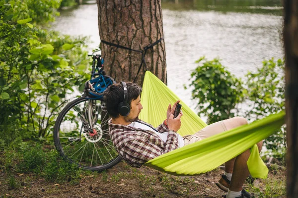 男は自転車で旅し 緑のハンモックでリラックスし スマートフォンでインターネットをサーフィンし 湖の近くの森の中でヘッドフォンで音楽を聴く 川沿いのキャンプ場でハンモックに身を包んだサイクリスト 男性オンバイクでハンモック — ストック写真