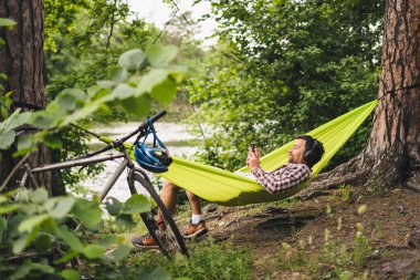 Adam bisikletle seyahat ediyor, yeşil hamakta dinleniyor, akıllı telefondan internette sörf yapıyor, gölün yanındaki ormanda kulaklıkla müzik dinliyor. Kamp alanındaki nehir kenarındaki hamakta bisikletçi. Hamakta bisikletli bir erkek..