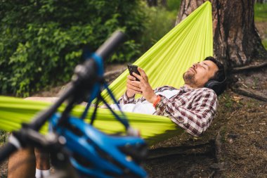 Genç beyaz adam hamakta dinleniyor, kulaklıkla müzik dinliyor ve sonrasında gölün yanındaki ormanda bisiklette akıllı telefon kullanıyor. Ses iyileştirme. Idyllic bir yer. Seyahat, doğada kamp..
