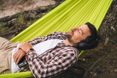 Göl kenarındaki kampta bisiklet süren bir adam müzik dinlerken yeşil bir hamakta dinleniyor. Doğada aktif eğlence teması. Kulaklıklı hippi bisikletçi nehir kenarındaki hamakta eğleniyor..