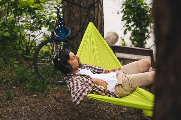 ハンモックに寝そべってヘッドフォンで音楽を聴いたり 湖の近くの森の中の自転車でスマートフォンを使用している若い白人男性 オーディオヒーリング 牧歌的な場所だ 自然の中でキャンプ — ストック写真