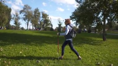 Kıdemli İskandinav kadın parkta izleme çubuklarıyla yürüyor. İskandinav yürüyüşlü yaşlı kadın dışarıda çalışıyor ve güneşli güzel bir günün tadını çıkarıyor. Etkin yaşam tarzı ve sağlık konsepti